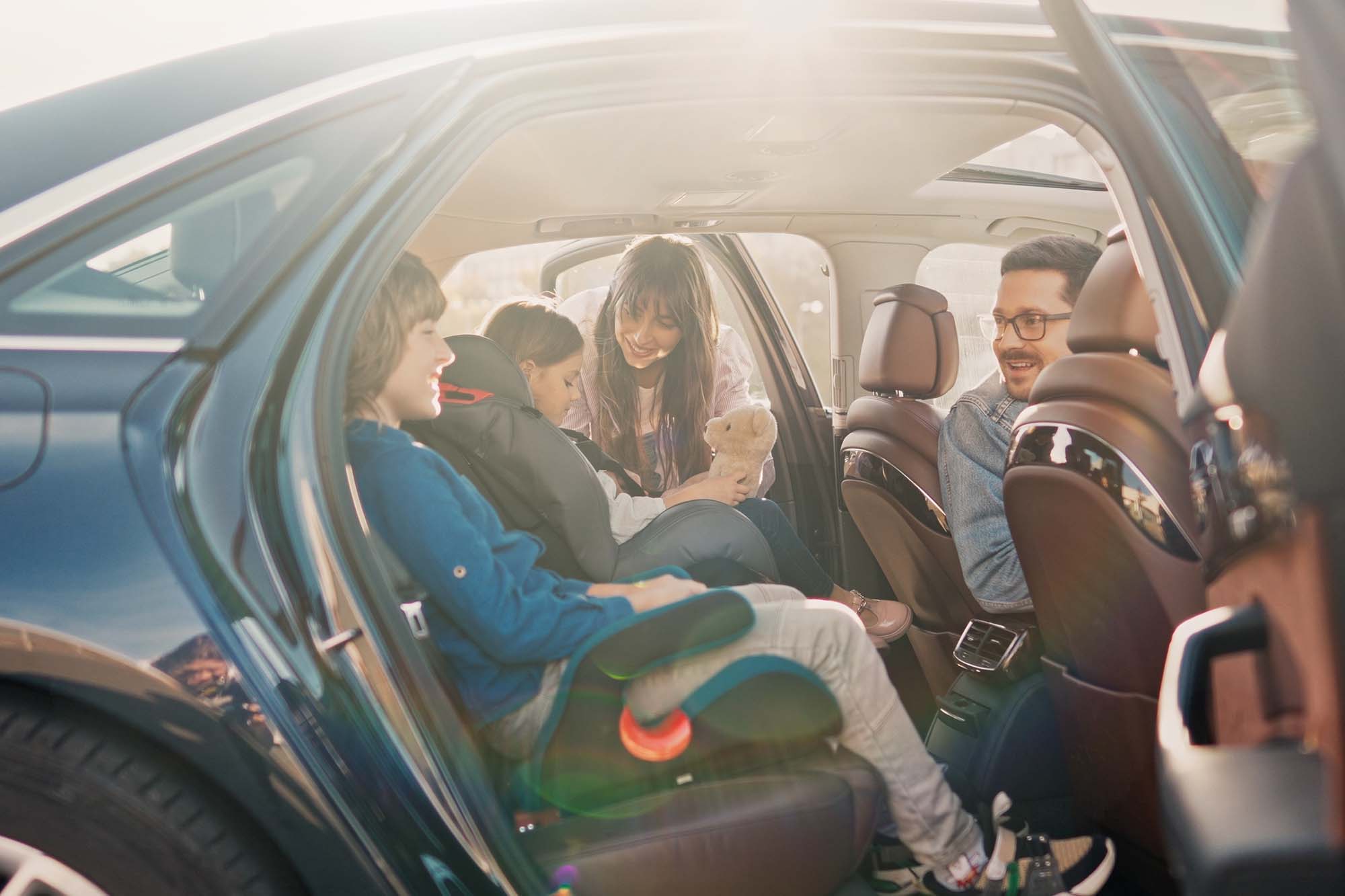 Family sitting inside a car with children in car seats and a woman holding a teddy bear, smiling and interacting.